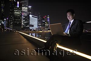Asia Images Group - Mature man working on a laptop at night, lit buildings as background