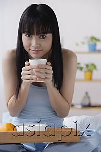 AsiaPix - Young woman sitting on bed, holding cup of coffee