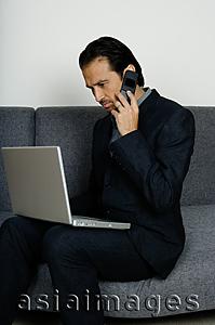 Asia Images Group - Businessman sitting on sofa, using mobile phone and looking at laptop