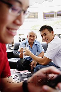 AsiaPix - Two men toasting with drinks, looking at camera, another man using mobile phone in the foreground